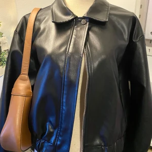 Svart skinnjacka med dragkedja - Snygg svart skinnjacka i mycket bra skick. Den har en stilren design med dragkedja och knappdetalj vid kragen. Perfekt för höst och vår, och funkar både till vardags och fest. Jackan har en normal passform och är långärmad.