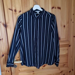 Svart och vit randig skjorta - Säljer en stilren mörkblå skjorta med vita ränder. Skjortan har långa ärmar och knappar framtill. Perfekt för både vardag och festliga tillfällen. Passar bra till både jeans och kostymbyxor!