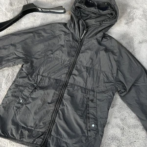 Svart vindjacka från C.P. Company - Säljer en svart vindjacka från C.P. Company med huva och inbyggda glasögon. Jackan har dragkedja framtill och praktiska fickor med knappar. Perfekt för blåsiga dagar och har en cool, unik stil.