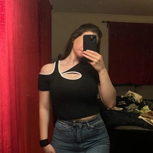 Svart offshoulder topp med unik design - Säljer en supercool svart offshoulder topp med en unik asymmetrisk design. Den har en snygg cut-out detalj framtill och vita kanter som ger en extra touch. Perfekt för en utekväll eller fest! Toppen är tight och croppad, vilket gör den perfekt att matcha med höga jeans eller kjol.