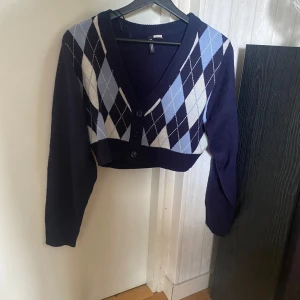 Mörkblå kofta med diamantmönster - Säljer en snygg mörkblå kofta med ett klassiskt diamantmönster i ljusblått och vitt. Koftan är croppad och har långa ärmar samt knappar framtill. Perfekt för höst och vår!