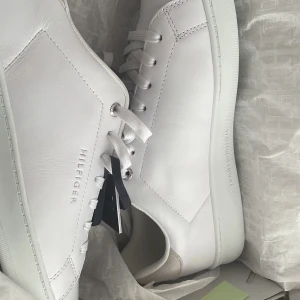 Vita sneakers från Tommy Hilfiger - Säljer ett par stilrena vita sneakers från Tommy Hilfiger. Skorna är i skinn och har en klassisk design med snörning. Perfekta för en casual look och passar till det mesta i garderoben. De är i nyskick och kommer med originalförpackning.