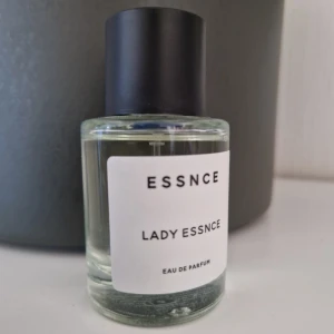 Lady Essnce Eau de Parfum - Säljer en elegant och fräsch parfymflaska från ESSNCE. Lady Essnce Eau de Parfum kommer i en stilren, genomskinlig glasflaska med en minimalistisk vit etikett och svart text. Flaskan har en svart kork som ger en modern touch. Perfekt för dagligt bruk eller speciella tillfällen.