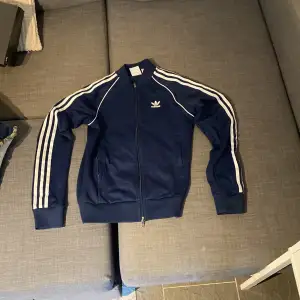 Säljer en snygg blå träningsjacka från Adidas med de klassiska tre ränderna längs ärmarna. Jackan har en dragkedja framtill och ribbade muddar vid ärmslut och nederkant. Perfekt för träning eller som en casual look.