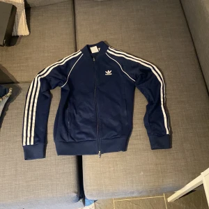 Blå träningsjacka från Adidas - Säljer en snygg blå träningsjacka från Adidas med de klassiska tre ränderna längs ärmarna. Jackan har en dragkedja framtill och ribbade muddar vid ärmslut och nederkant. Perfekt för träning eller som en casual look.