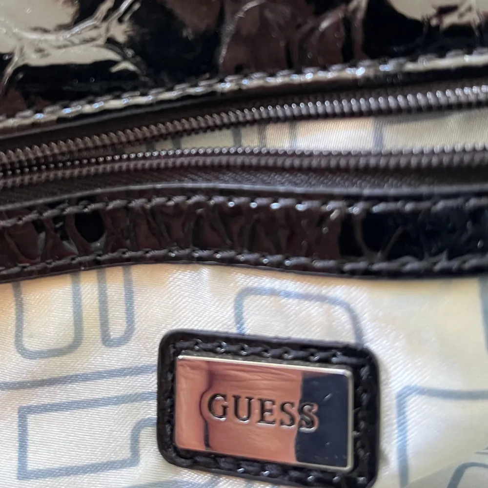 Guess handväska i flerfärgat tyg med ”guess” tryckt  på. Läderliknande handtag och på kant runt lock. Silvertonad hårdvara med ”Guess” präglat. Invändig blixtlåsficka och en extra liten ficka  inuti. Silkesliknande tyg på insidan. Magnetisk stängning. Väskor.