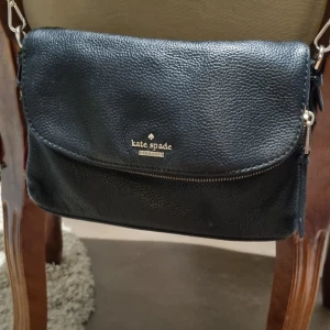 Svart axelväska från Kate Spade - Säljer en snygg svart axelväska från Kate Spade i skinnimitation. Väskan har en praktisk dragkedja som går att ta bort och ett stilrent randigt foder inuti. Den har även en yttre del med två fack i. Perfekt för både vardag och fest!