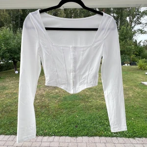 Vit croppad topp - En vit croppad topp med långa ärmar. Toppen har en öppning/stängning mitt på framsidan i form av hyskor och hakar 💞 Säljer pga att den aldrig blev använd!  (ALDRIG ANVÄND)  Längd: 41 cm Axel: 36 cm Byst: 79 cm Ärmlängd: 61 cm