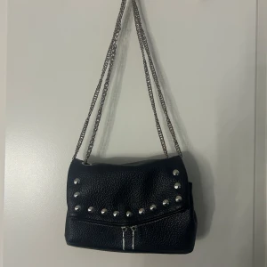 Svart axelväska från bershka - Snygg svart axelväska i syntetmaterial. Väskan har en kedja som axelrem och en dragkedja för säker förvaring. 