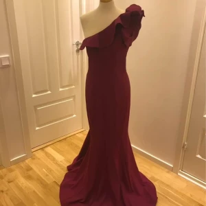 Vinröd axelbandslös klänning - Elegant vinröd/burghundy långklänning med en offshoulder-design och volangdetaljer. Säljes för 1500kr, storlek 40-42 (går att sy in)