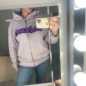  Doganci kofta  - Säljer en ljuslila hoodie från Doganci med dragkedja och en cool lila text på bröstet. Perfekt för en avslappnad stil. Den har en bekväm passform och en praktisk huva.