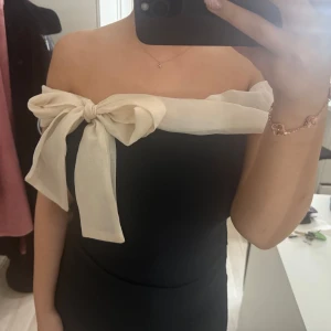 Svart offshoulder body med rosett - Elegant svart offshoulder body med en stor beige rosett över bysten. Perfekt för en stilren look med en touch av elegans. Bodyn har en tight passform som framhäver figuren.🖤