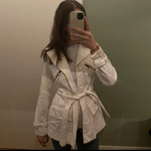 Vit manchester trenchcoat - Snygg vit trenchcoat i manchester med knappar och bälte i midjan. Jackan har långa ärmar och dragkedjor på bröstfickorna för en cool look. Perfekt för att hålla stilen under kyligare dagar.
