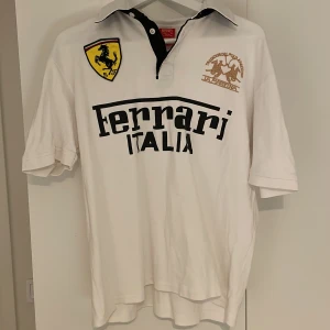 Vit kortärmad Ferrari piké  - Säljer en vit kortärmad t-shirt från Ferrari med svart text 'Ferrari Italia' på framsidan. T-shirten har en broderad Ferrari-logga och en annan broderad detalj i guld. Perfekt för bilentusiaster!