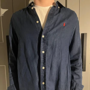 Mörkblå skjorta från Ralph Lauren - Säljer en stilren mörkblå skjorta från Ralph Lauren med klassisk krage och knappar framtill. Skjortan har en broderad logotyp i rött på bröstet och långa ärmar. 