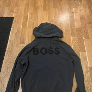 Grå hoodie från Boss - Säljer en stilren svart hoodie från Boss med stor logga på ryggen. Tröjan har en klassisk design med huva och långa ärmar. Perfekt för en avslappnad look.