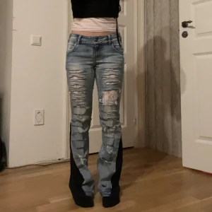 Blå slitna jeans med svarta detaljer - Säljer ett par blå jeans med slitna detaljer och svarta inslag på benen. De har en bootcut-stil och är lågmidjade. Perfekta för en trendig look med en twist.
