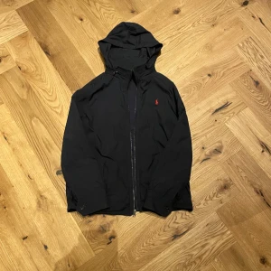 Polo Ralph Lauren vindjacka - En riktigt snygg vindjacka från Polo Ralph Lauren. Den är flisfodrad och relativt tunn perfekt till våren. Storleken är 14-16/ Xs och passar från 164-174 beroende på hur man vill att den ska sitta. Väldigt bra skick och inga defekter. Luvan är även infällbar.