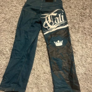 Blå jeans med tryck från Bsat - Unika blå jeans från Bsat med stort vitt tryck 'Cali' och en krona på ena benet. Byxorna har en klassisk jeanslook med en modern twist. Perfekta för dig som vill sticka ut med stil.