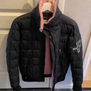 Svart quiltad jacka från Pelle Pelle - Snygg svart quiltad jacka från Pelle Pelle med rosa foder. Jackan har en hög krage och dragkedja framtill. Den har långa ärmar och en broderad logga på ärmen. Perfekt för kyligare dagar!