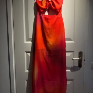 Färgglad långklänning med knytband - Säljer en färgglad långklänning i orange och rosa toner. Klänningen har smala axelband och en öppen rygg med knytband. Perfekt för sommarens varma dagar.