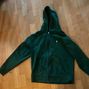 Grön hoodie från Polo Ralph Lauren - Säljer en snygg grön hoodie från Polo Ralph Lauren. Skicket är bra, inga skador eller andra defekter. Dock en liten fett fläck på framsidan, men den synds inte så länge man inte kollar noga. Storleken är L men passar även M då den är lite kort i armarna för mig. Pris kan diskuteras vid snabb affär.