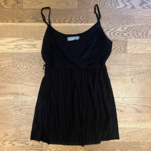 Svart omlottklänning från Lindex - Snygg svart omlottklänning från Lindex med smala axelband och plisserad design. Perfekt för en stilren look. Klänningen har en v-ringad framdel och är tillverkad i ett lätt och bekvämt material.