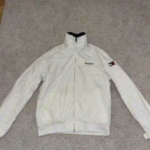 Vit jacka från Tommy Hilfiger - Säljer en stilren vit jacka från Tommy Hilfiger med dragkedja och broderad logga på ärmen. Jackan har en hög krage med Hilfiger-tryck och är perfekt för kyligare dagar. Den är långärmad och har en klassisk passform.