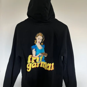 Svart hoodie från Revenite - Säljer en svart hoodie från Revenite med en cool tryckdesign på baksidan som föreställer en kvinna i blå klänning och texten 'fru gårman'. Hoodien har en känguruficka och snörning vid huvan. Perfekt för en avslappnad stil.
