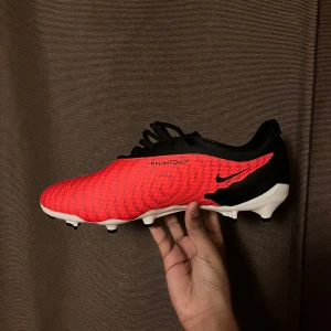 Nike Phantom GT2 fotbollsskor - Snygga Nike Phantom GT2 fotbollsskor i rött och svart med snörning. Perfekta för fotbollsspelare. Boxen finns inte men garanterat att du får den i en box