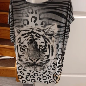 Grå tunika med tigertryck från Jassire - Säljer en cool grå tunika från Jassire med ett stort tigertryck framtill. Tunikan har korta ärmar och ett unikt mönster med ränder och fläckar i svart och grått. Perfekt för dig som vill sticka ut!