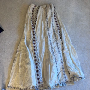 Vit långkjol med broderade detaljer - Säljer en vacker vit långkjol med broderade blommor i rött och svart. Kjolen har en luftig passform och elastisk midja för extra komfort. Perfekt för en bohemisk stil! Den är inte struken så ni tänker på det annars är den rakare. Säljer pågrund av flytt och för mycket kläder 😄😄