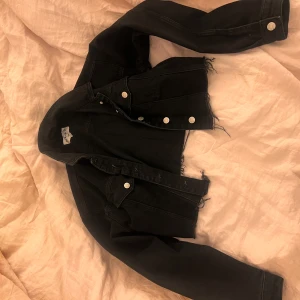 Svart croppad jeansjacka - Snygg svart croppad jeansjacka från NA-KD med fransiga kanter och silverfärgade knappar. Perfekt för en trendig look. Jackan har långa ärmar och klassisk krage.