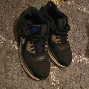 Svarta Nike sneakers - Säljer ett par svarta Nike sneakers med snörning och blå detaljer. Skorna har en klassisk design med en bekväm passform och är perfekta för vardagsbruk.