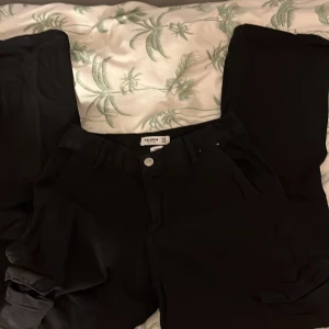 Svarta cargopants från Pull&Bear - Snygga svarta cargopants från Pull&Bear med praktiska fickor på sidorna. Byxorna har en modern design och är perfekta för en avslappnad stil. De har en knapp och dragkedja framtill.