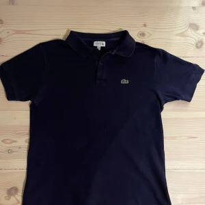 Mörkblå pikétröja från Lacoste - Säljer en klassisk mörkblå pikétröja från Lacoste med korta ärmar och den ikoniska krokodilloggan på bröstet. Perfekt för en stilren och avslappnad look. Tröjan har en krage med knappar framtill.
