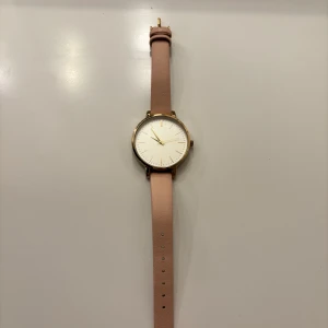 Elegant klocka med rosa armband - Stilren klocka med ett mjukt rosa skinnarmband och en vit urtavla från H&M. Klockan har en guldig boett och spänne, vilket ger en lyxig känsla. Perfekt accessoar för att lyfta din stil.