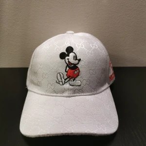 Vit keps med Musse Pigg  - Snygg vit keps från ggi med Musse Pigg-broderi på framsidan. Kepsen har ett diskret mönster av gg-loggor och texten gg' på sidan. Perfekt för att ge en touch av lyx och lekfullhet till din stil.