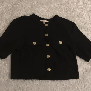 Svart kavaj med guldknappar - Snygg svart kavaj med guldknappar framtill och två fickor på bröstet. Kavajen har en croppad passform och trekvartsärmar, perfekt för en stilren look.