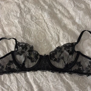 Svart spets-bh från Nelly - Elegant svart bh från Nelly med vacker spetsdetaljering och blommönster. Den har justerbara axelband och en klassisk hakknäppning bak. Perfekt för att känna sig extra fin. Bh:n sälj pågrund av att jag aldrig använt den. I storlek 75B.