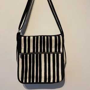 Randig axelväska från Marimekko - Snygg axelväska från Marimekko med svartvita ränder. Väskan har en praktisk dragkedja och en justerbar axelrem. Perfekt för att bära med sig det nödvändigaste med stil. Bra skick!
