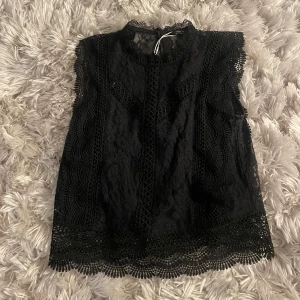 Svart spetsblus - Säljer en elegant svart spetsblus med vackra detaljer och spetsig rygg. Blusen är ärmlös och har en fin spetskant vid ärmslut och nederkant. 