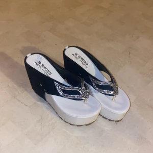 Svarta sandaler med kilklack och strassdetaljer - Snygga svarta sandaler med hög kilklack och glittriga strassdetaljer på remmarna. Sandalerna har en vit platåsula som ger extra höjd och komfort. Perfekta för att ge en elegant touch till din outfit.