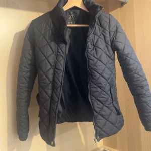 Mörkblå quiltad vårjacka  - Snygg  quiltad jacka med dragkedja och knappar. Jackan har en klassisk design med långa ärmar och en hög krage