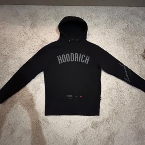 Svart hoodie från Hoodrich - Snygg svart hoodie från Hoodrich med tryck på bröstet och ärmen. Den har en klassisk passform med långa ärmar och en bekväm huva. Skicka 8/10, storlek XS men passar alla som har S, lite sliten vid höger armbåge.  Priset kan diskuteras! 
