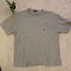 Grå t-shirt från Polo Ralph Lauren - Säljer en klassisk grå t-shirt från Polo Ralph Lauren i 100% bomull. T-shirten har en bröstficka med den ikoniska loggan broderad. Perfekt för en avslappnad stil och i perfekt skick.