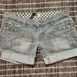 Grå jeansshorts från August Jeans - Snygga grå jeansshorts från August Jeans med slitna detaljer och fransade kanter. Shortsen har flera fickor, en med dragkedja och broderade ord på baksidan. Perfekta för en avslappnad stil/sthlms stil! 💗💗 39cm rakt över, 29 cm från höft till ben.