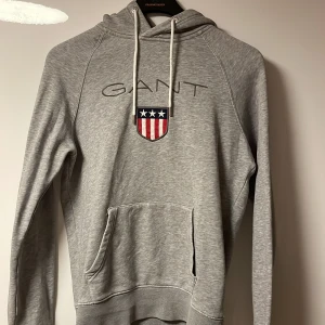 Grå hoodie från GANT - Säljer en grå hoodie från GANT med logotyp och emblem på bröstet. Tröjan har en justerbar huva med snören och en praktisk magficka. Perfekt för en avslappnad stil. Bara att höra av dig om du har några frågor!