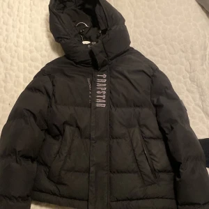 Svart pufferjacka från Trapstar - Säljer en svart pufferjacka från Trapstar med stor logga på ryggen och textdetaljer på framsidan. Jackan har en huva och dragkedja framtill. Perfekt för kyliga dagar. Vill ni ha fler bilder skriv privat. 