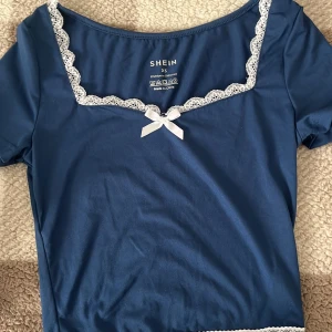 Blå topp med spetsdetaljer från Shein - Säljer en söt blå topp från Shein i storlek XS. Toppen har korta ärmar och är dekorerad med vit spets längs halsringningen samt en liten rosett framtill. Materialet är en mjuk blandning av polyester och elastan.